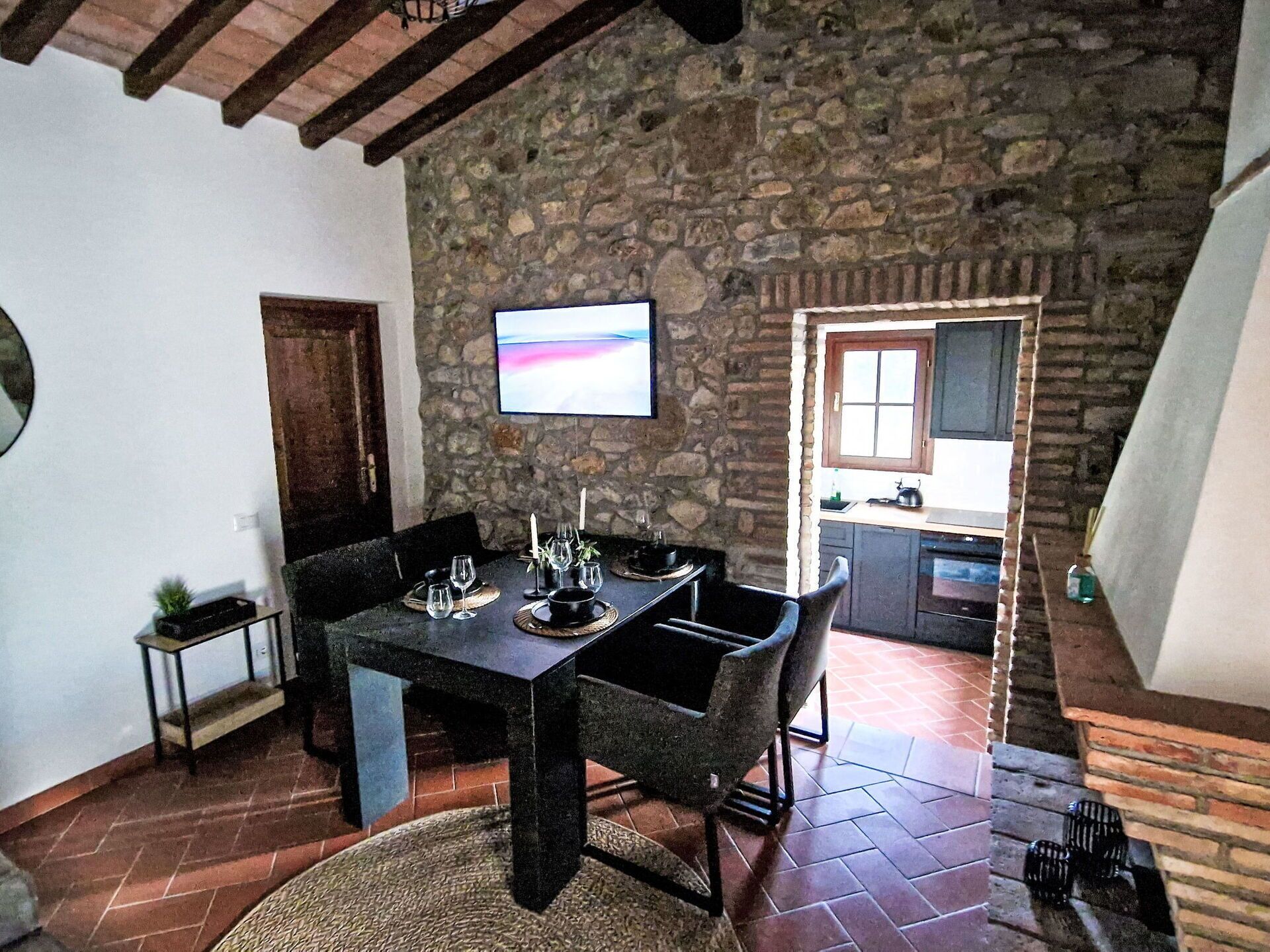 A dining area with a stone wall, a TV mounted above the table, and a kitchen visible through an open door.