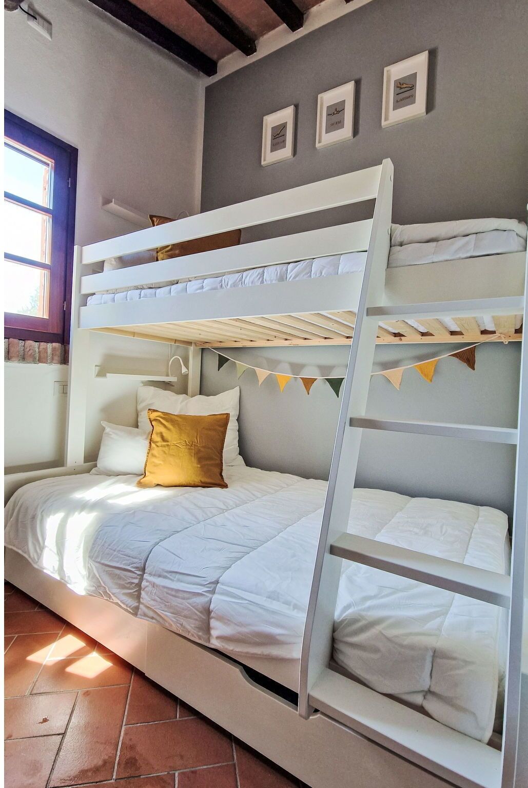 Een stapelbed met een ladder, wit beddengoed en een geel kussen. Er hangen fotolijstjes aan de muur en een raam met uitzicht.
