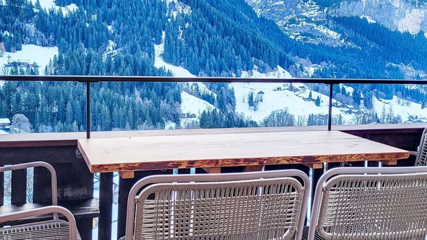 Une terrasse avec une table en bois et des chaises en osier, offrant une vue sur un paysage montagneux enneigé.