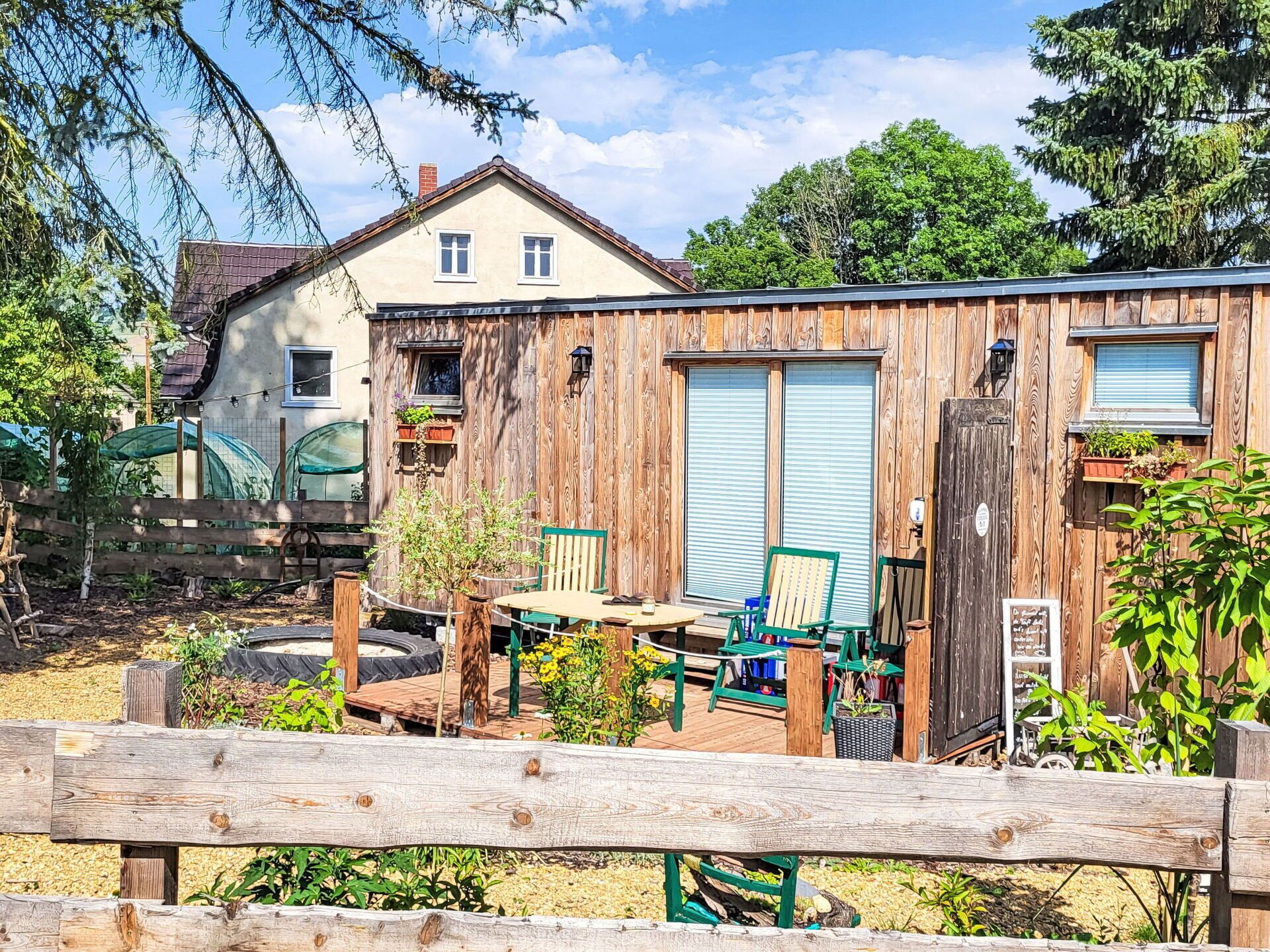 Een rustiek houten gebouw met een garagepoort, een kleine tafel met stoelen, en een tuin met planten en een houten hek.