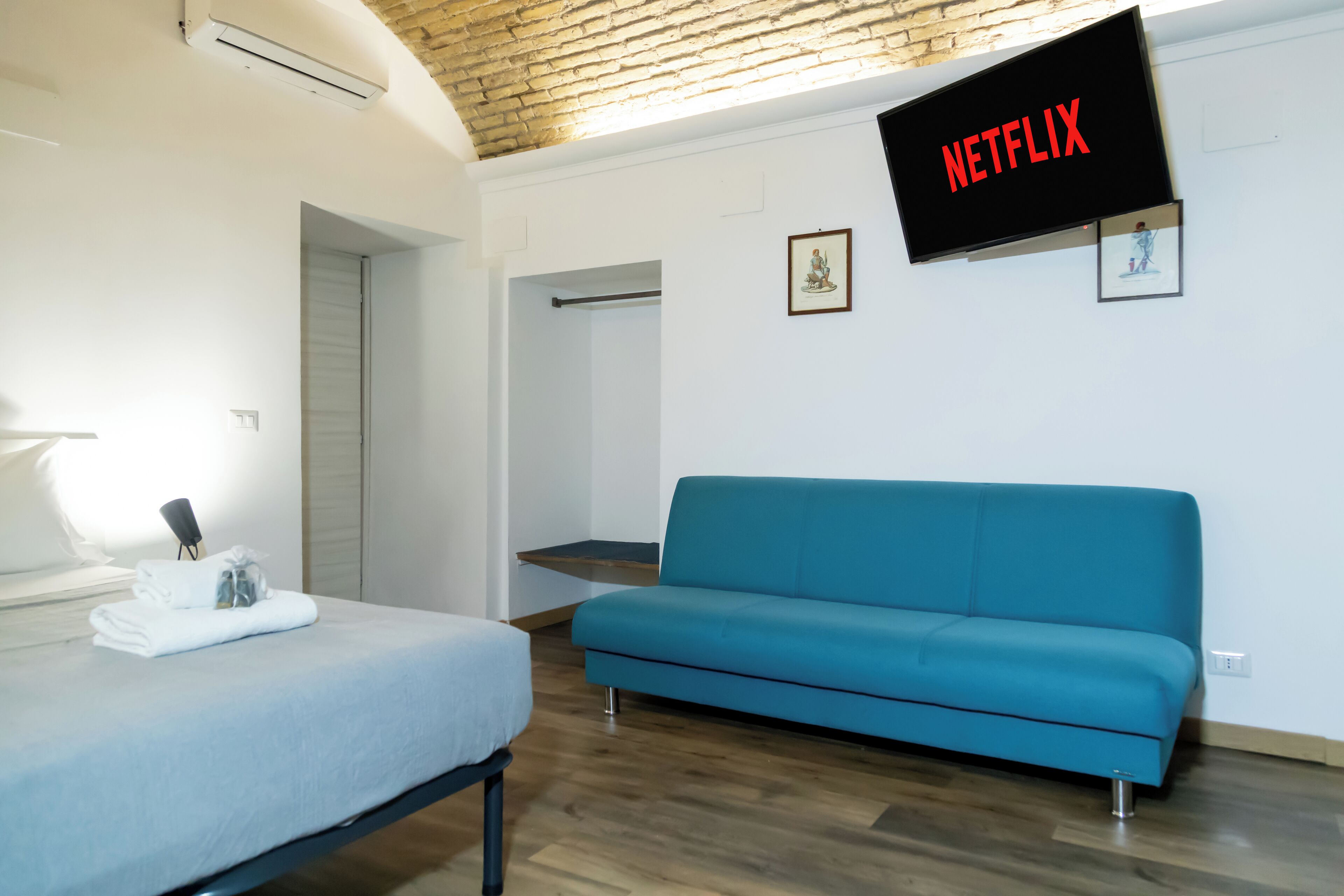 A room with a bed, a blue sofa, a wall-mounted TV displaying 'NETFLIX', and a wooden ceiling.