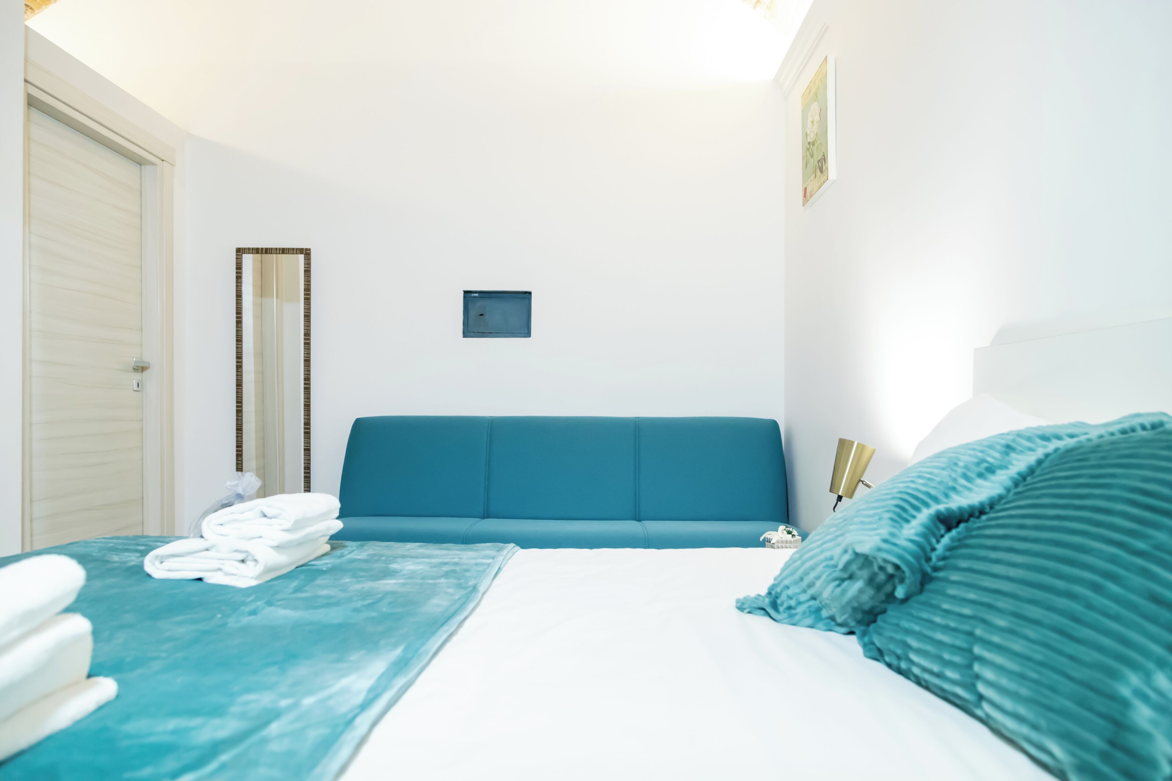 A hotel room with a bed, a blue sofa, a small table with a lamp, and a wall-mounted mirror.