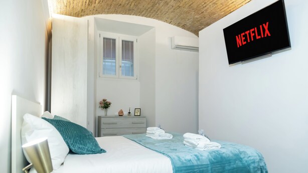 A modern bedroom with a bed, a bedside table, a lamp, a wall-mounted TV displaying "NETFLIX", and a window with blinds. - Termini Volte Antiche & Comfort (Rome)