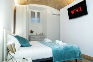 Ein modernes Schlafzimmer mit einem Bett, einem Nachttisch, einer Lampe, einem am Wand montierten Fernseher, der "NETFLIX" anzeigt, und einem Fenster mit Jalousien.