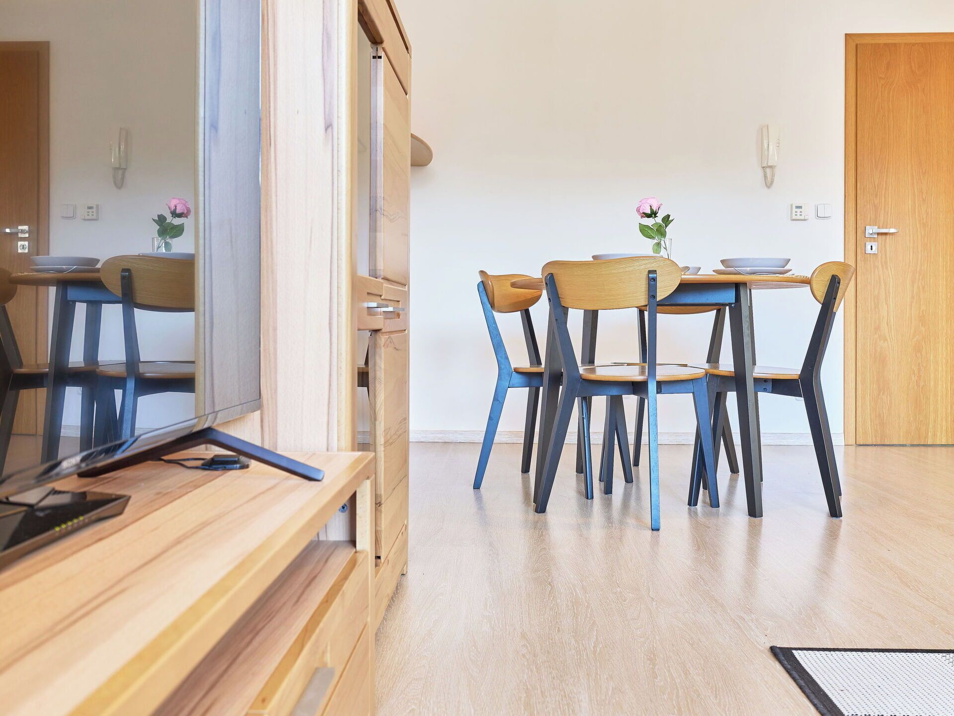 A dining area with a wooden table and chairs, a mirror, and a small vase with a flower.