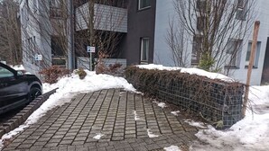En asfalterad gångväg med snöfläckar, en parkerad bil och en byggnad med ett stängsel av metall och snöhögar.
