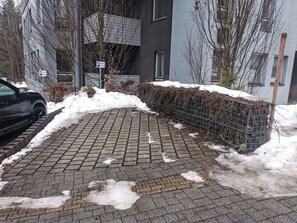 Ein gepflasterter Weg mit Schneeflecken, ein abgestelltes Auto und ein Gebäude mit einem Metallzaun und Schneebedeckung.