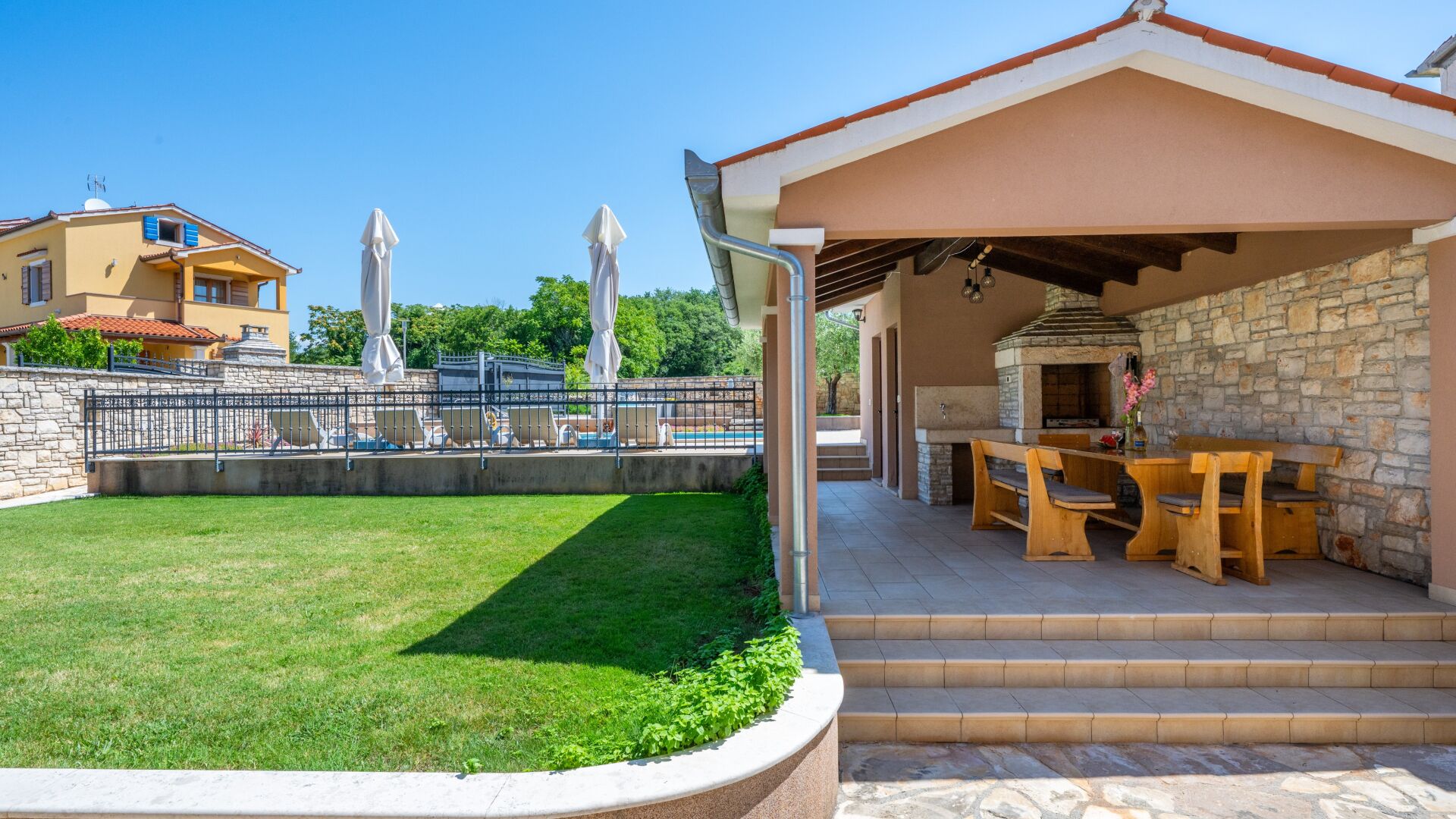 A covered outdoor area with a barbecue and seating, a stone wall, and a pool in the background.