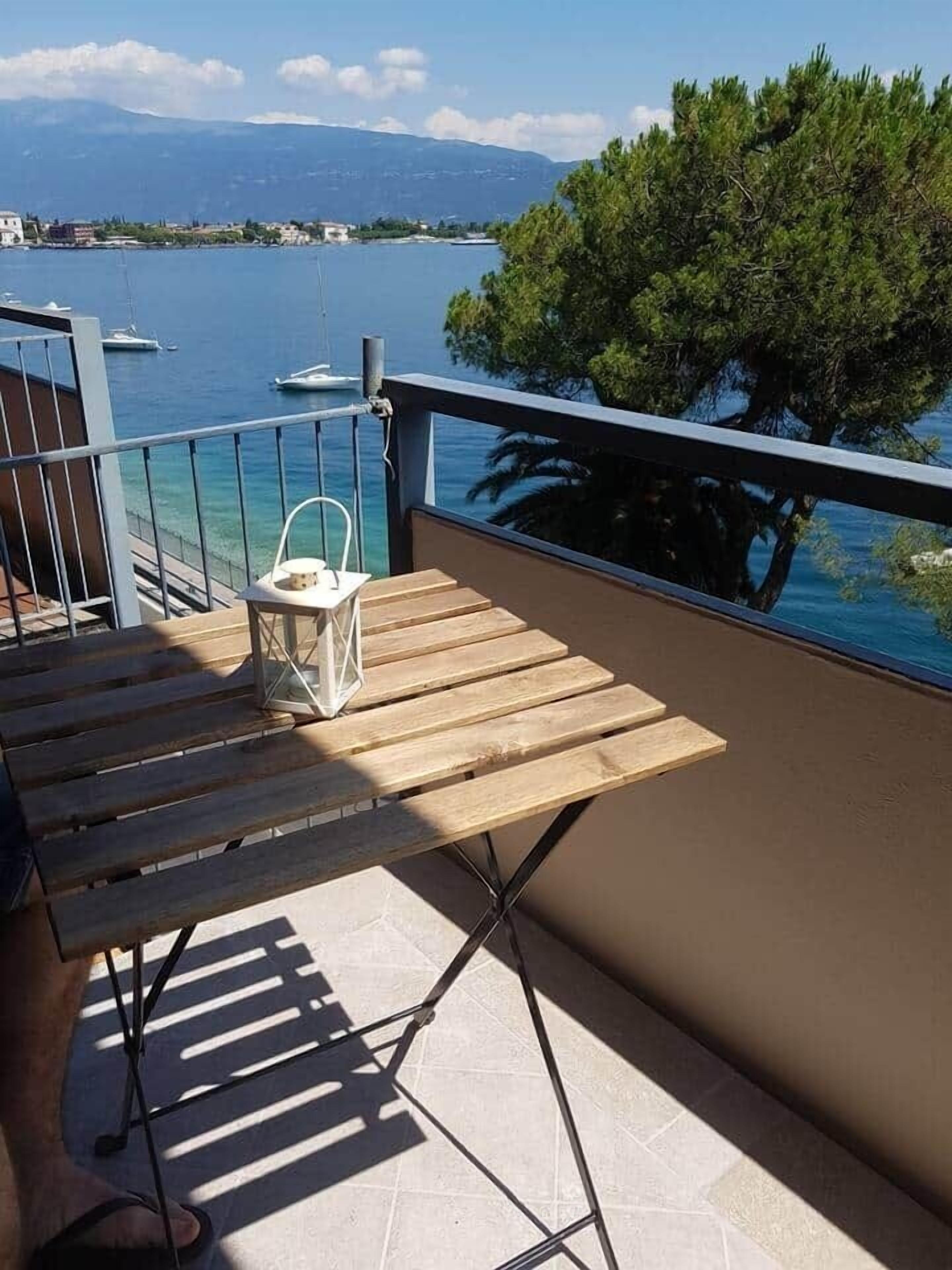 Ein Balkon mit Holztisch und Stühlen mit Blick auf ein Gewässer, auf dem Boote fahren, und einer fernen Küste.