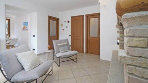 Ein Zimmer mit einem grauen Sofa, einem grauen Sessel und einem kleinen Tisch mit einem Weinglas. Zwei Holztüren und eine Ziegelwand sind ebenfalls zu sehen.