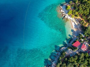 Eine Vogelperspektive einer Küstenlandschaft mit klarem türkisblauem Wasser, einem Sandstrand und einer Ansammlung von Gebäuden mit roten Dächern.