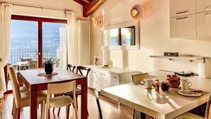 Zona pranzo con tavolo e sedie in legno, vista mare attraverso ampie finestre e cucina con elettrodomestici moderni.