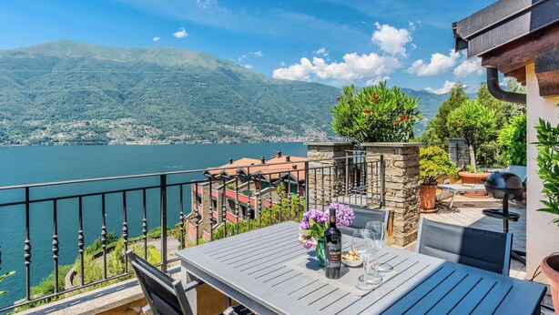 Una terrazza con tavolo, sedie, una bottiglia e bicchieri, con vista su un lago e montagne.