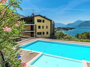 Ein Pool mit Blick auf einen See und Berge, ein gelbes Gebäude mit Balkonen und einem klaren Himmel.