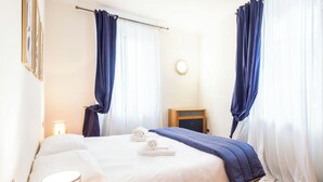 Ein ordentlich bezogenes Bett mit weißen Leinen und einer blauen Decke, eine Nachttischlampe und ein Nachttisch aus Holz mit einer Lampe.