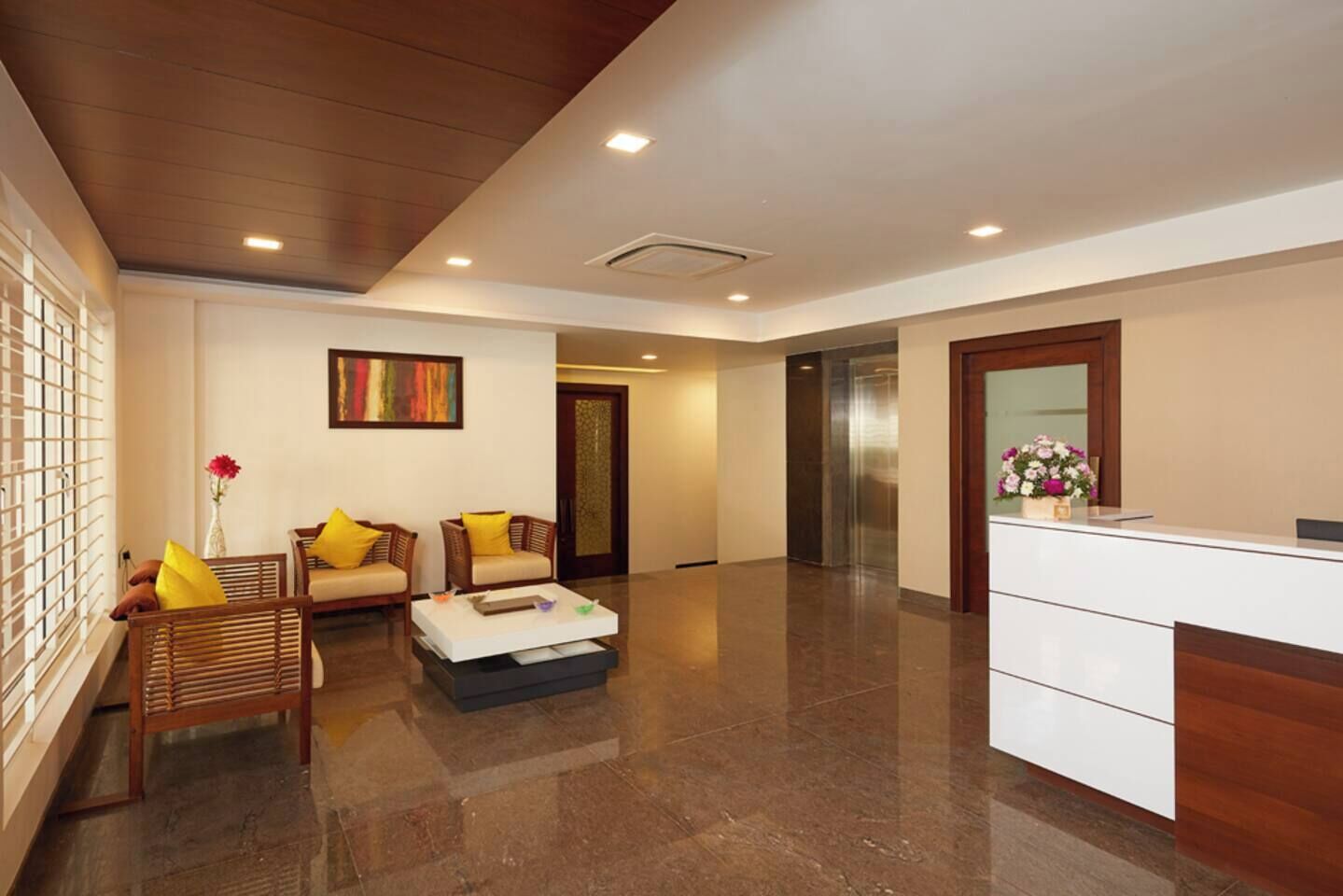 A spacious lobby with a reception desk, two sofas with yellow cushions, a coffee table, and a large painting on the wall.
