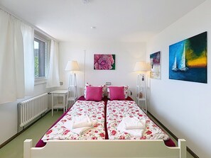 Un lit double avec une literie à motifs floraux, deux lampes de chevet, un radiateur, une fenêtre avec des rideaux et deux tableaux accrochés au mur.