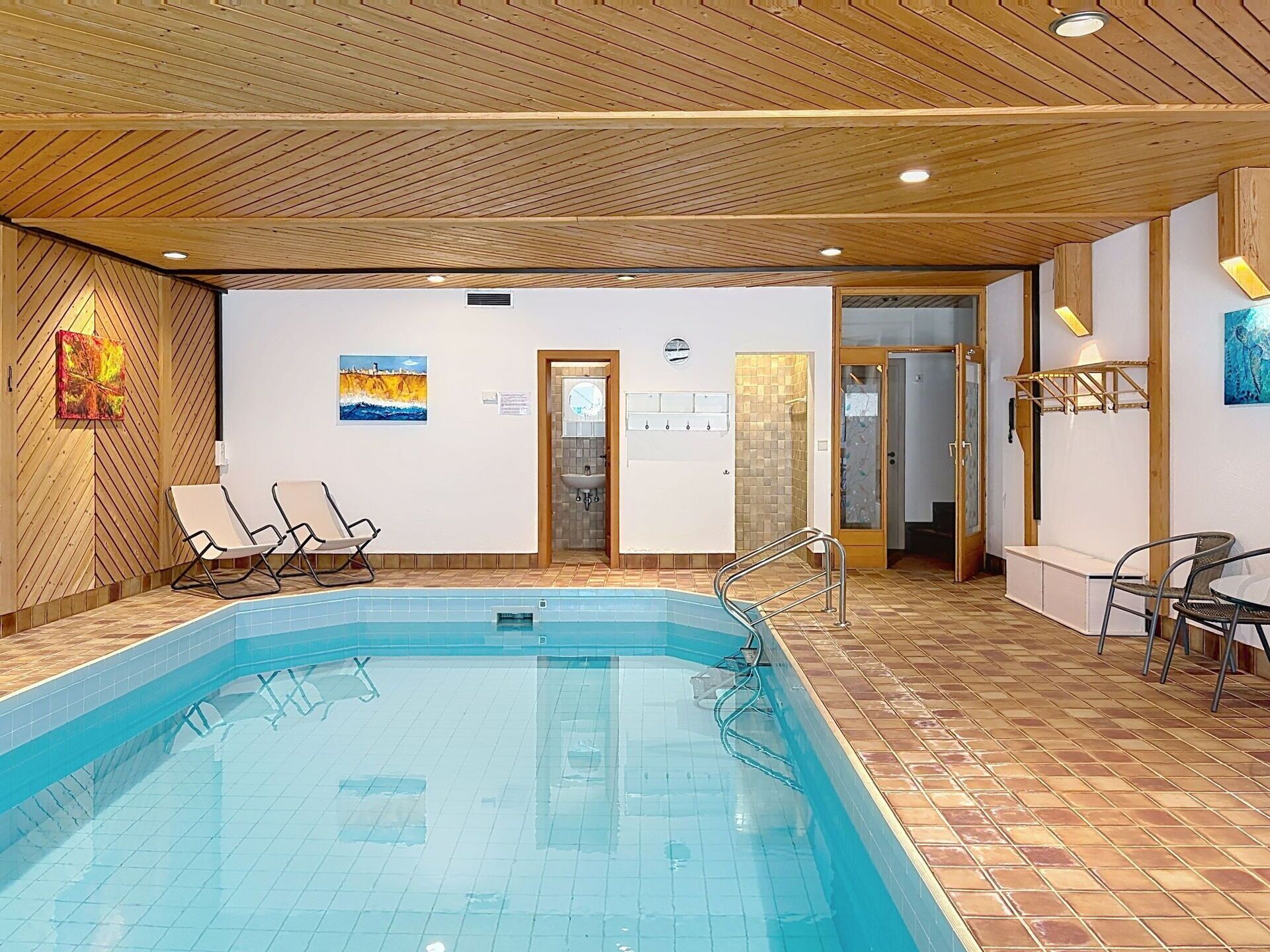 An indoor swimming pool with tiled flooring, a wooden ceiling, and a door leading to a shower area.
