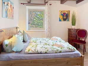 Ein Schlafzimmer mit einem Holzbett, einem fenster mit Vorhängen, einem roten Sessel, einer Holzkommode und zwei Bildern an der Wand.
