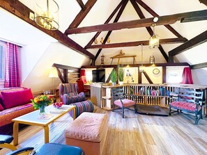 Een gezellig loft-achtig vertrek met zichtbare houten balken, een open haard, een boekenkast en een comfortabele zithoek met een bank en stoelen.