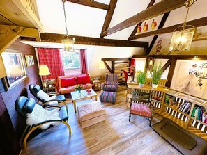 Un salon spacieux avec un plafond haut, des poutres en bois, un canapé rouge, des fauteuils à rayures, une table basse et une bibliothèque.