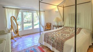Ein Himmelbett mit gemustertem Überwurf, ein stehender Spiegel, ein Nachttisch mit Lampe sowie ein Blick ins Freie durch eine Glastür.