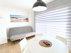 Ein Zimmer mit einem weißen runden Tisch, einem grauen Sofa, einer schwarzen Pendelleuchte und Blick auf den Strand durch Jalousien.