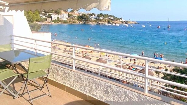 Un balcón con mesa y sillas, con vista a una playa donde se ven gente y barcos.