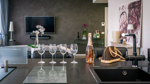 A modern hotel room with a flat-screen TV, a bar area with wine glasses, a bottle of champagne, and a Louis Vuitton bag on the counter. - Rosa 37 - Modern & Elegamnt 2BR Apt - Near Venice (Venezia)