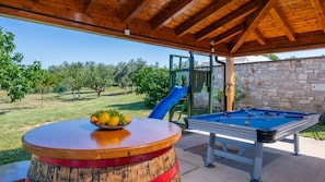 A pool table, a fruit bowl, and a slide in a covered outdoor area with a stone wall and trees in the background. - Villa Milena Vodnjan by Villas Guide (Vodnjan)