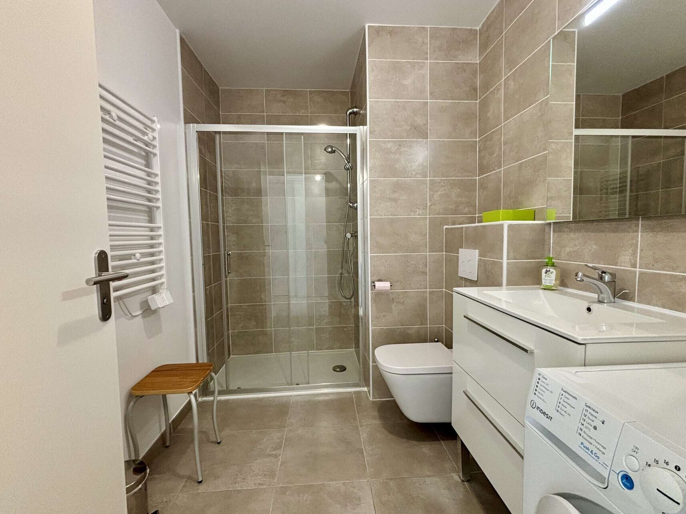Une salle de bain moderne avec une douche à parois de verre, des toilettes, un lavabo avec un miroir et un petit tabouret en bois.