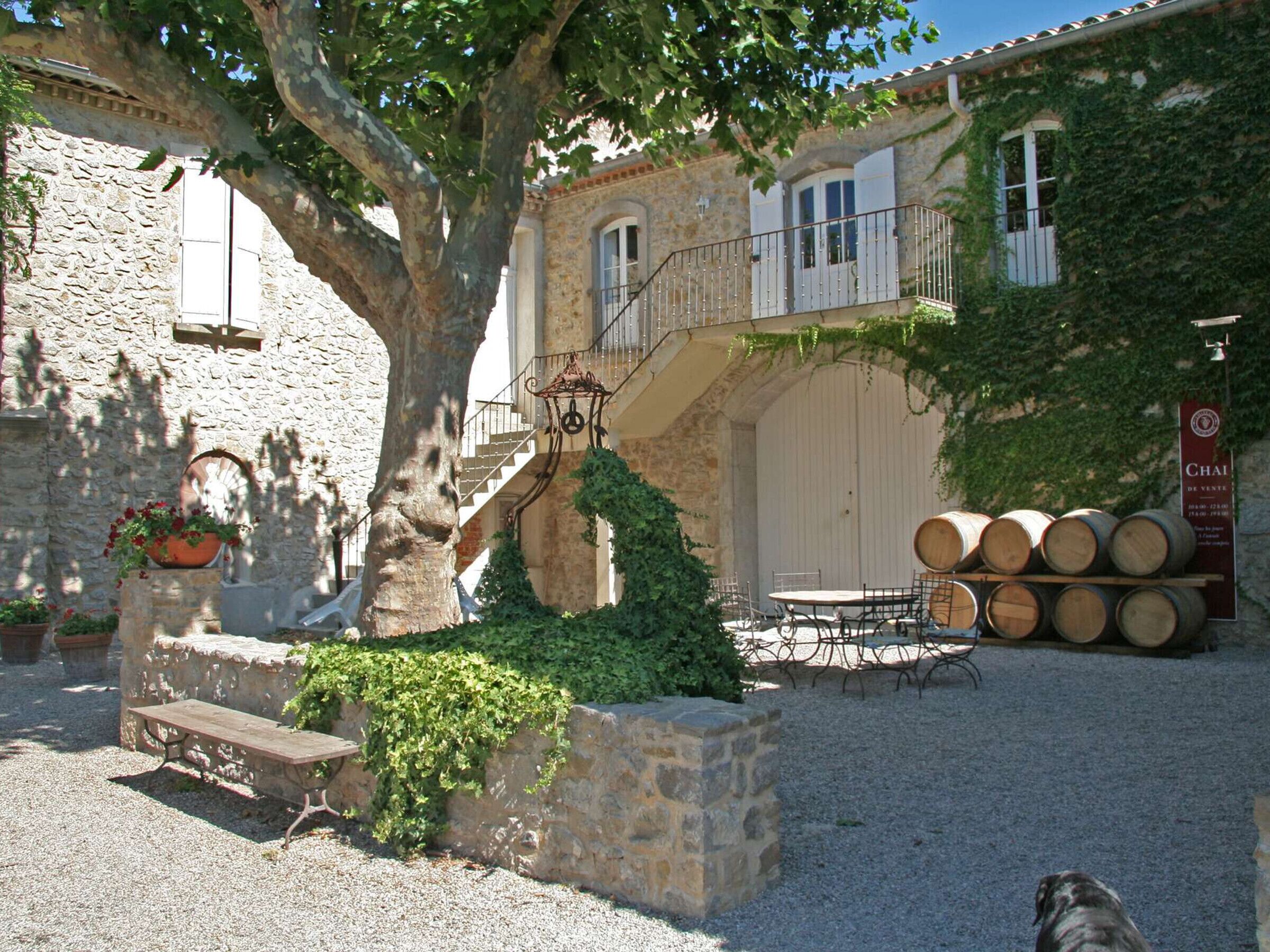 A courtyard with a stone bench, a table, and barrels, surrounded by stone walls and a tree.