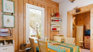 Une pièce chaleureuse, avec des murs en bois, une table à manger, des chaises en osier, un buffet blanc avec des tiroirs et une étagère où sont disposés de la vaisselle et des verres.