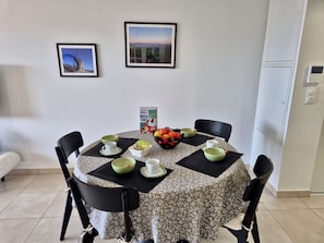 A dining area with a round table covered by a patterned tablecloth, surrounded by black chairs. There are bowls, cups, and a box of frosted flakes on the table. Two framed pictures are hung on the wall above the table. - 3-Room Apartment in Cogolin, 4 Pers with Parking Included (Cogolin)