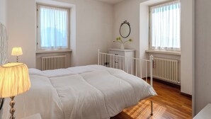 Quarto com cama, abajur, cômoda, espelho e duas janelas com cortinas.