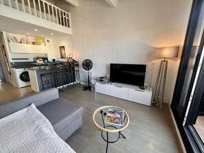 A modern living room with a grey sofa, a round coffee table with a magazine, a TV on a white cabinet, a fan, and a kitchen area with a washing machine and a dining table. - Studio 4 Pers. Terrace in Marines de Cogolin (Cogolin)