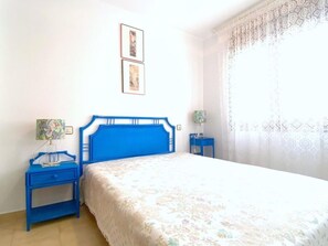 Ein Schlafzimmer mit blauem Kopfteil, einem Bett mit floraler Bettdecke, zwei Nachttischen mit Lampen und einem Fenster mit gemusterten Vorhängen.