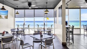 Un restaurant doté d’une terrasse offrant une vue sur la plage, avec des chaises et des tables en métal, de grandes fenêtres et un ventilateur de plafond.