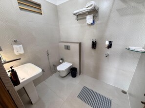 A modern bathroom with a white sink, toilet, and shower. There are towels and toiletries on the wall. - Sharnam On The Ganges (Rishikesh)