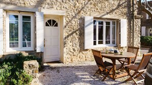 A stone building with a white door and windows, a gravel patio, and a wooden table with chairs. - Charming 2-Bedroom Cottage in Vineyard Estate, Near Narbonne-Plage, Sleeps 5, with WiFi and Parking (Narbonne-Narbonne Plage-Narbonne Plage)