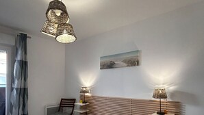 Ein Schlafzimmer mit Bett, Stuhl, Nachttischen, Lampe, einem Bild an der Wand und einem Fenster mit Vorhängen.