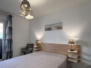 Ein Schlafzimmer mit Bett, Stuhl, Nachttischen, Lampe, einem Bild an der Wand und einem Fenster mit Vorhängen.