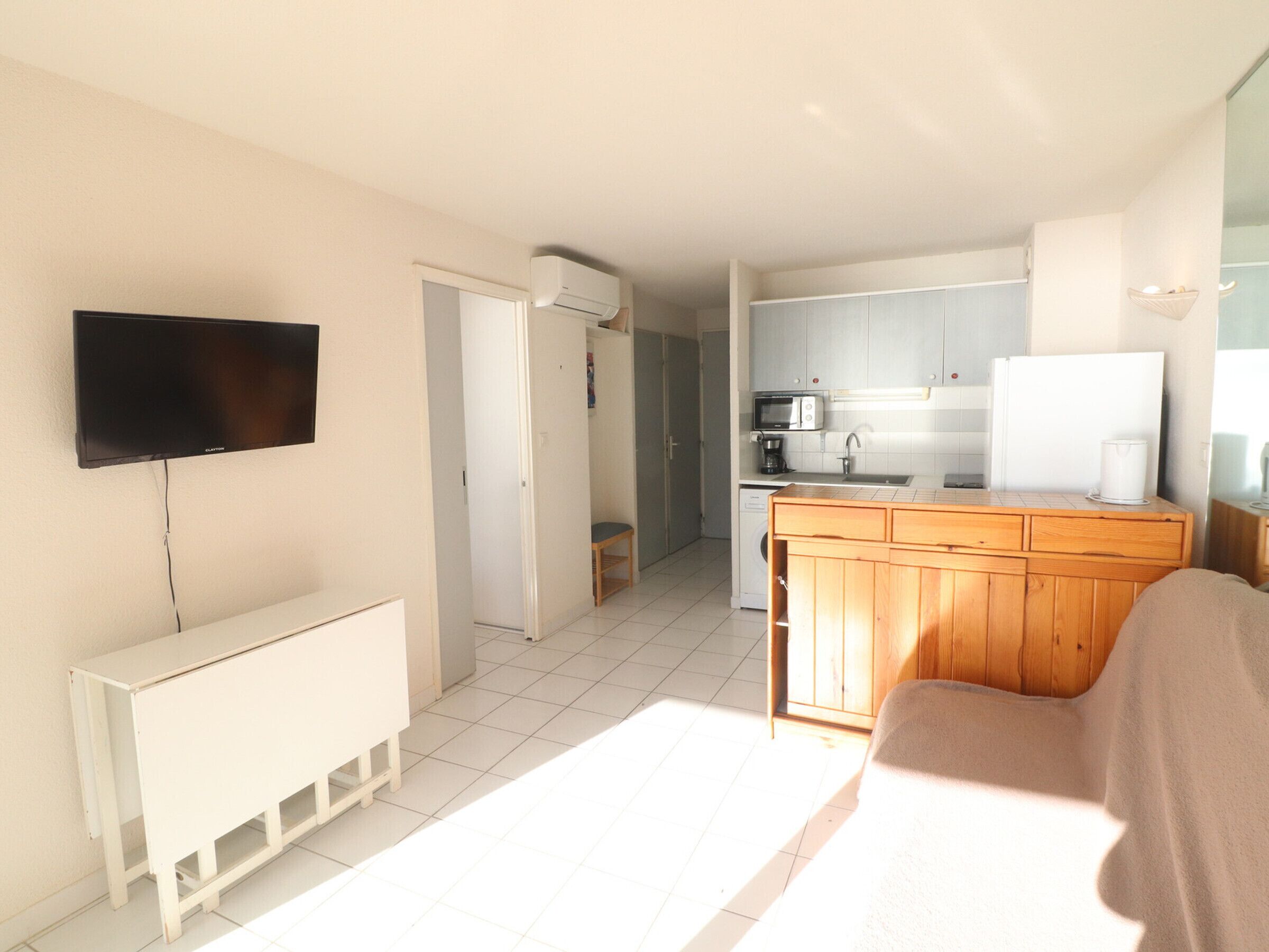 A compact living space with a kitchenette, a television mounted on the wall, and a small seating area.