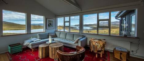 A spacious living room with a large window overlooking a natural landscape, a grey sofa, a wooden coffee table, and a rustic chair.