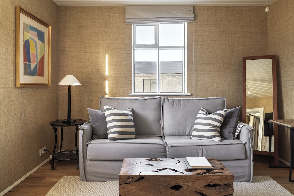 Una sala de estar moderna con un sofá gris, almohadones a rayas, una mesa de centro de madera, una lámpara de pie y un amplio ventanal con persianas.