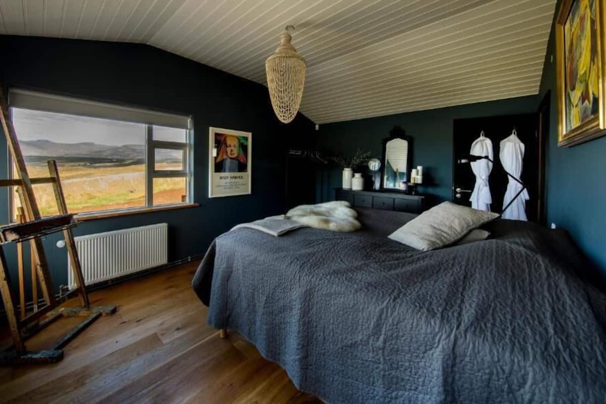 Un dormitorio con una cama grande, una escalera de madera, un radiador, una ventana con vistas al campo y una araña.