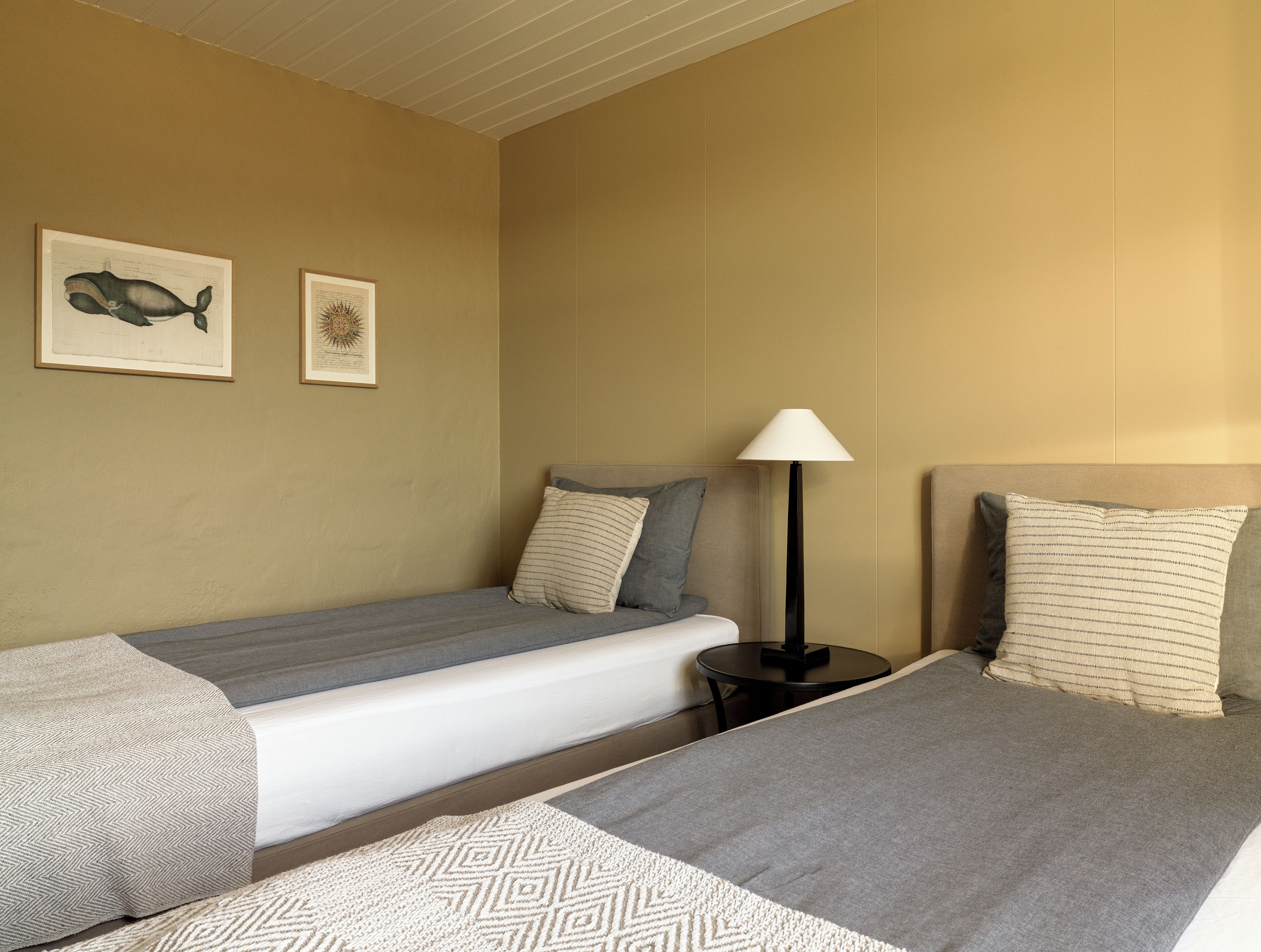A hotel room with two single beds, a nightstand, a lamp, and two framed pictures on the wall.