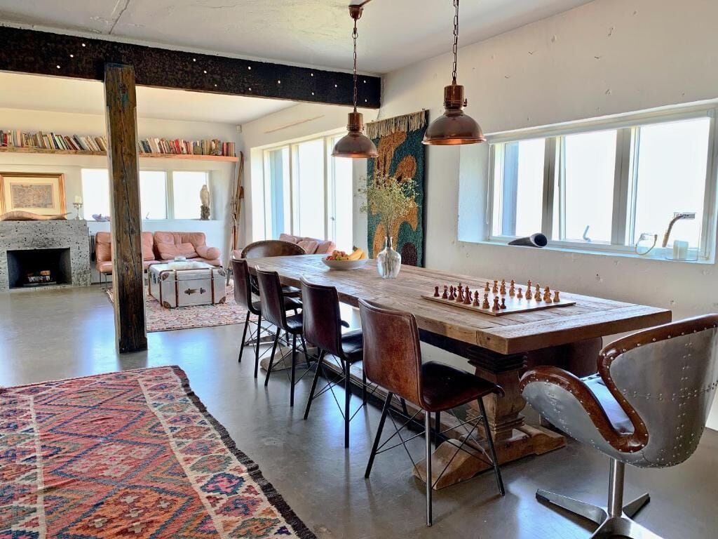 A spacious living area with a large wooden dining table, multiple chairs, and a chessboard. There is a fireplace, bookshelves, and a large window providing natural light.