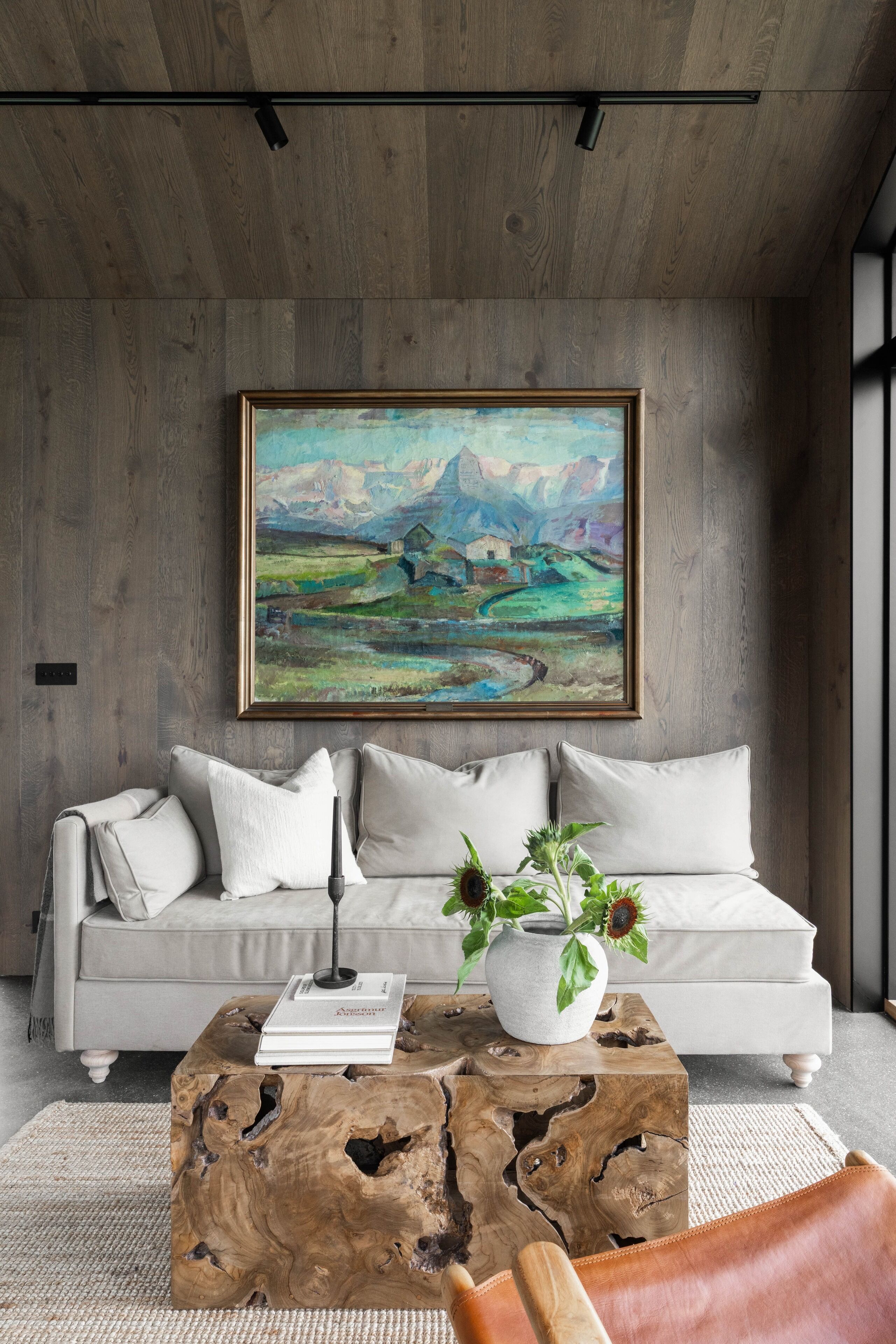 Una sala moderna con un sofá gris, una mesa de centro de madera con un libro y un jarrón de flores, y un cuadro de un paisaje en la pared.
