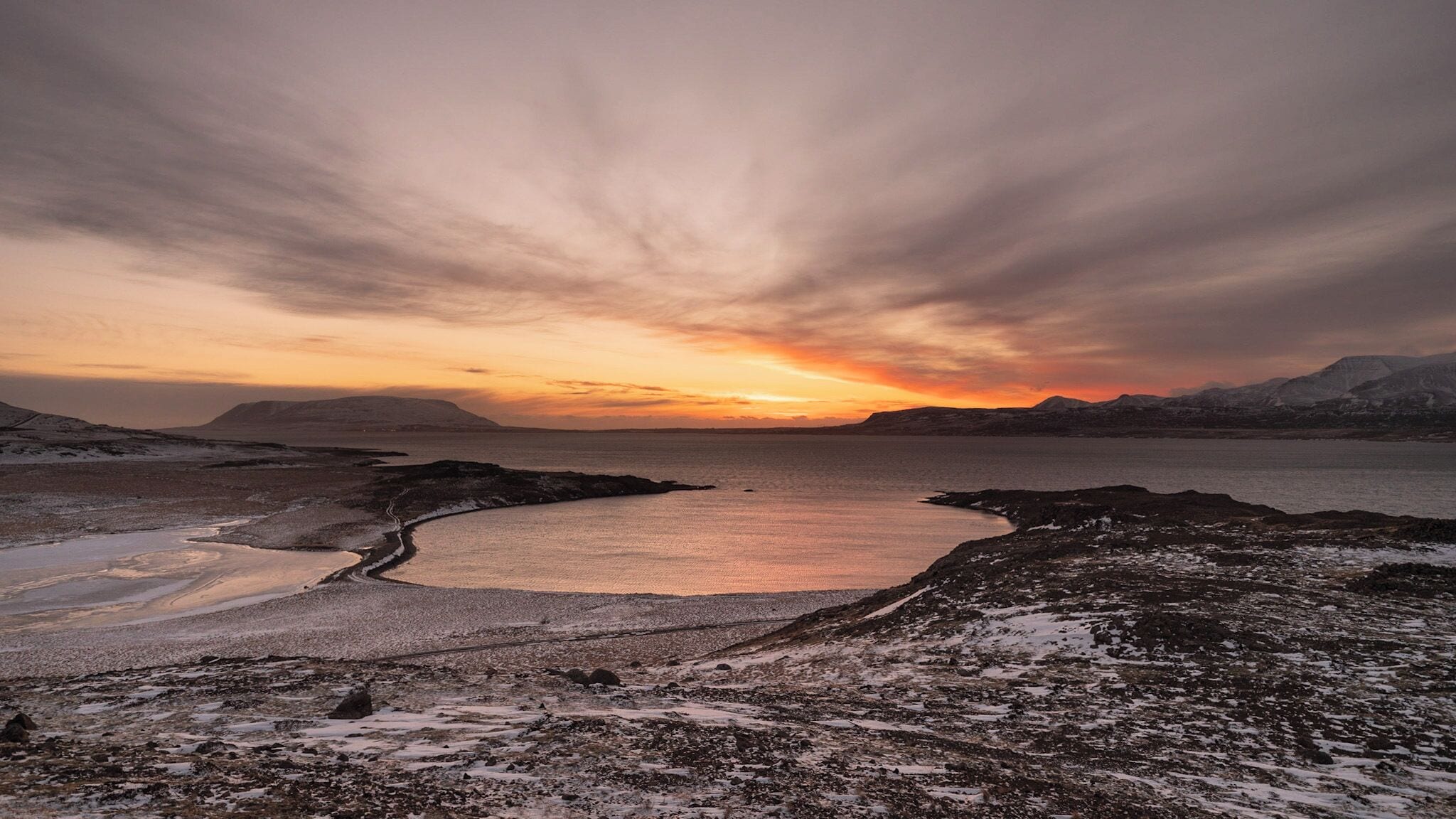 A coastal landscape with a frozen lake, snow-covered terrain, and distant mountains under a sunset sky.